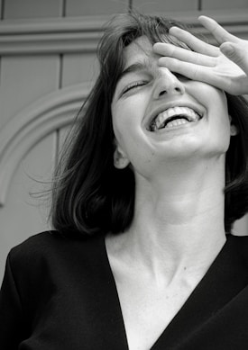 A monochrome photograph featuring a young woman with a joyful expression. Her head is tilted slightly back, her mouth open in laughter, and her eyes partially covered by her hand. The image captures a candid, carefree moment.