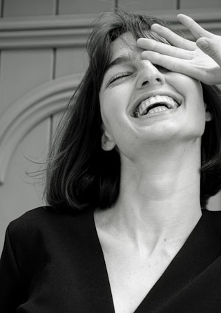 A monochrome photograph featuring a young woman with a joyful expression. Her head is tilted slightly back, her mouth open in laughter, and her eyes partially covered by her hand. The image captures a candid, carefree moment.