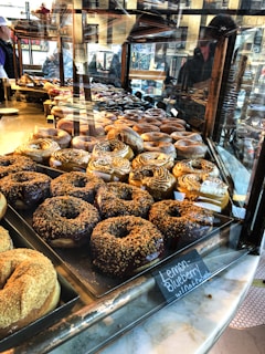 A display case filled with an assortment of donuts of various flavors and toppings, including cinnamon swirls and chocolate-coated ones. Reflections from the glass reveal the bustling activity of a city street, with people visible outside.