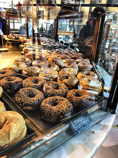 A display case filled with an assortment of donuts of various flavors and toppings, including cinnamon swirls and chocolate-coated ones. Reflections from the glass reveal the bustling activity of a city street, with people visible outside.