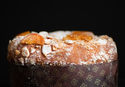 A close-up view of a baked dessert with a dusting of powdered sugar and almond slices on top. The surface has an appealing golden-brown texture, and there are pieces of candied fruit adding color and texture. The dessert is wrapped with decorative paper.