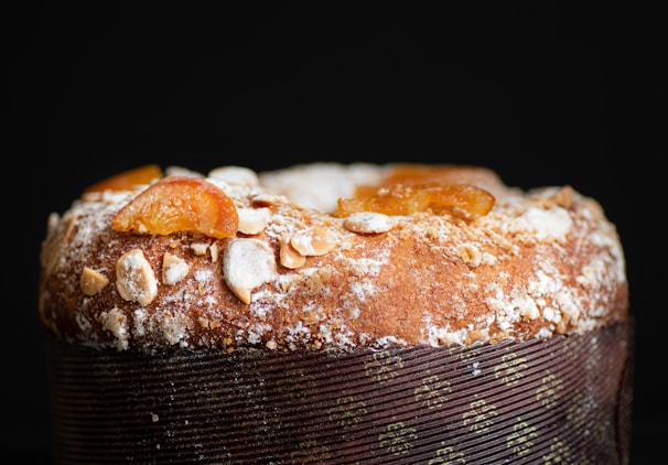A close-up view of a baked dessert with a dusting of powdered sugar and almond slices on top. The surface has an appealing golden-brown texture, and there are pieces of candied fruit adding color and texture. The dessert is wrapped with decorative paper.