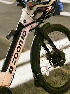 A close-up of the front part of a bicycle with the brand name 'Zoomo' visible on the frame. A helmet is placed on the handlebar, and the scene suggests it's taken on a street possibly at night or in low light conditions.