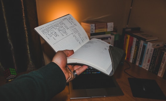A hand holding an open notebook with handwritten notes. The scene is set on a desk with several books lined up against the wall. A soft ambient light illuminates the area, casting a warm glow. A Rubik's Cube and a closed laptop are also visible on the desk.