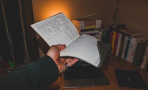A hand holding an open notebook with handwritten notes. The scene is set on a desk with several books lined up against the wall. A soft ambient light illuminates the area, casting a warm glow. A Rubik's Cube and a closed laptop are also visible on the desk.