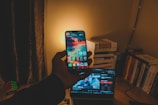 A hand holds a smartphone displaying a colorful home screen with apps. The background reveals a dimly lit room with a stack of books, a laptop showing a video, and a Rubik's Cube on a wooden desk. Warm, yellowish light illuminates the scene.