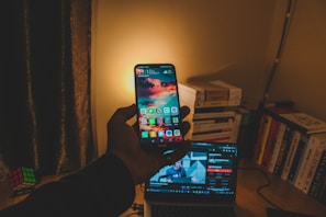 A hand holds a smartphone displaying a colorful home screen with apps. The background reveals a dimly lit room with a stack of books, a laptop showing a video, and a Rubik's Cube on a wooden desk. Warm, yellowish light illuminates the scene.