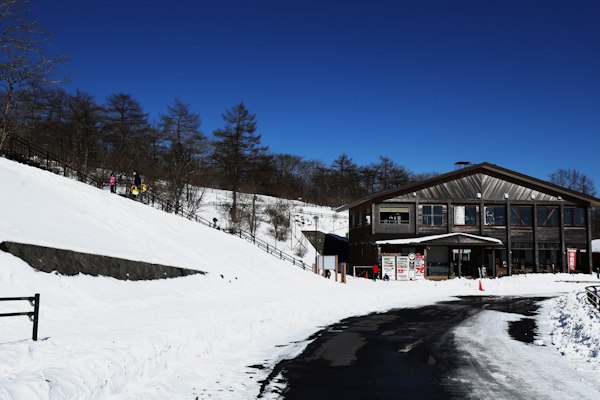 A timber base lodge beside a snowy slope, with a freshly plowed road in front and skiers walking up the hill