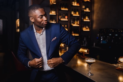 A well-dressed man in a navy suit stands confidently at a dimly lit bar, adjusting his jacket. A cocktail in a tall glass sits on the sleek counter. Shelves filled with bottles are illuminated in the background, adding an elegant ambiance.