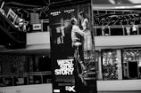 A large poster of the movie West Side Story is displayed prominently inside a shopping mall. The poster features a black and white image of a man and a woman in a romantic pose, positioned at the center. Surrounding the poster are mall balconies and shopfronts, with visible details such as sale signs and decorated railings.