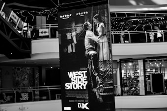 A large poster of the movie West Side Story is displayed prominently inside a shopping mall. The poster features a black and white image of a man and a woman in a romantic pose, positioned at the center. Surrounding the poster are mall balconies and shopfronts, with visible details such as sale signs and decorated railings.