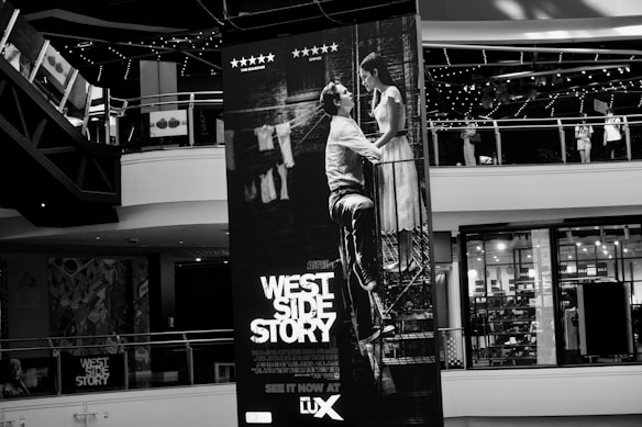 A large poster of the movie West Side Story is displayed prominently inside a shopping mall. The poster features a black and white image of a man and a woman in a romantic pose, positioned at the center. Surrounding the poster are mall balconies and shopfronts, with visible details such as sale signs and decorated railings.
