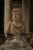 A marble bust of a bearded man with curly hair, draped in a toga, is displayed prominently in an ornately decorated hall. The sculpture is set on a pedestal with an inscription plaque at the base. It is surrounded by large marble columns, contributing to a grandiose and stately atmosphere.