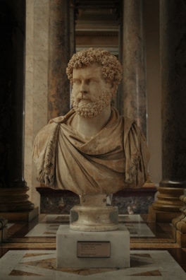 A marble bust of a bearded man with curly hair, draped in a toga, is displayed prominently in an ornately decorated hall. The sculpture is set on a pedestal with an inscription plaque at the base. It is surrounded by large marble columns, contributing to a grandiose and stately atmosphere.