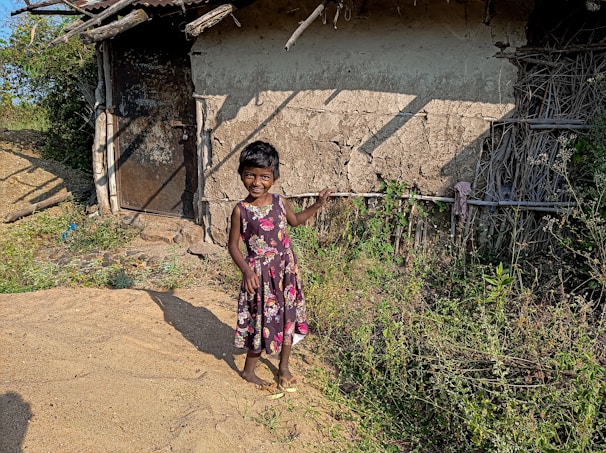 A young girl proudly holding her school books with a big smile in front of a modest home.