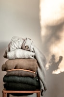 A neatly stacked pile of variously colored knitted sweaters sits on a wooden chair. Each sweater displays a different knit pattern and texture, ranging from light to dark shades. Soft natural light casts a shadow on the wall, adding warmth to the scene.