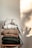 A neatly stacked pile of variously colored knitted sweaters sits on a wooden chair. Each sweater displays a different knit pattern and texture, ranging from light to dark shades. Soft natural light casts a shadow on the wall, adding warmth to the scene.