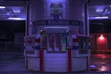 A token purchasing machine area at night, illuminated by blue and purple lights. The area is tiled with white squares and consists of several token vending machines, a bill changer, and various signs, including one that reads 'TOKENS PURCHASED HERE'. The ground is wet and there are red support poles around the machines.