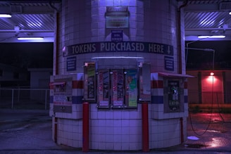 A token purchasing machine area at night, illuminated by blue and purple lights. The area is tiled with white squares and consists of several token vending machines, a bill changer, and various signs, including one that reads 'TOKENS PURCHASED HERE'. The ground is wet and there are red support poles around the machines.