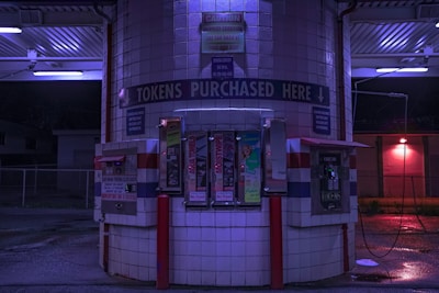 A token purchasing machine area at night, illuminated by blue and purple lights. The area is tiled with white squares and consists of several token vending machines, a bill changer, and various signs, including one that reads 'TOKENS PURCHASED HERE'. The ground is wet and there are red support poles around the machines.