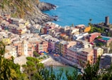 A vibrant coastal town with an array of colorful buildings perched along a cliffside overlooking the blue sea. The architecture is nestled between rocky cliffs and lush greenery, with charming streets and an ancient tower visible. The waterfront is lively with people enjoying the beach, and umbrellas dot the sandy shore.