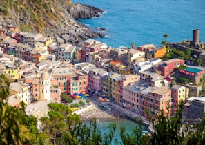 A vibrant coastal town with an array of colorful buildings perched along a cliffside overlooking the blue sea. The architecture is nestled between rocky cliffs and lush greenery, with charming streets and an ancient tower visible. The waterfront is lively with people enjoying the beach, and umbrellas dot the sandy shore.