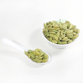A white ceramic spoon and a white ceramic bowl are filled with green cardamom pods. The background is a plain, light surface.
