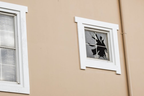 A beige wall with two windows, one of which is intact and the other has a shattered glass pane. The frames around the windows are white, and the shattered window reveals cracks and missing pieces.