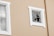 A beige wall with two windows, one of which is intact and the other has a shattered glass pane. The frames around the windows are white, and the shattered window reveals cracks and missing pieces.