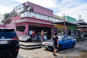 A damaged building with a pink exterior is partially destroyed, with debris scattered around. People are standing and discussing the situation, while a security guard observes. Two parked cars, one black and one blue, are visible in the foreground. The scene conveys a sense of aftermath and recovery.
