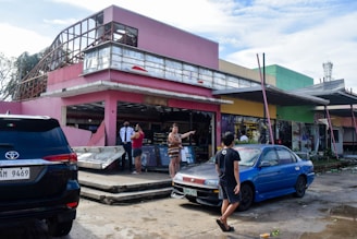 A damaged building with a pink exterior is partially destroyed, with debris scattered around. People are standing and discussing the situation, while a security guard observes. Two parked cars, one black and one blue, are visible in the foreground. The scene conveys a sense of aftermath and recovery.