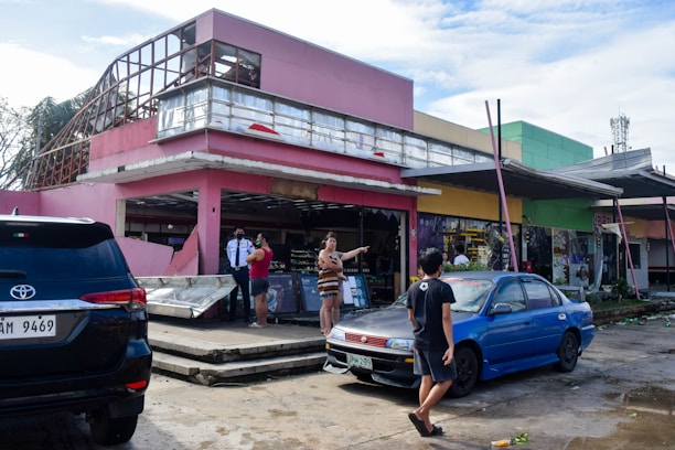 A damaged building with a pink exterior is partially destroyed, with debris scattered around. People are standing and discussing the situation, while a security guard observes. Two parked cars, one black and one blue, are visible in the foreground. The scene conveys a sense of aftermath and recovery.