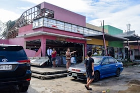 A damaged building with a pink exterior is partially destroyed, with debris scattered around. People are standing and discussing the situation, while a security guard observes. Two parked cars, one black and one blue, are visible in the foreground. The scene conveys a sense of aftermath and recovery.