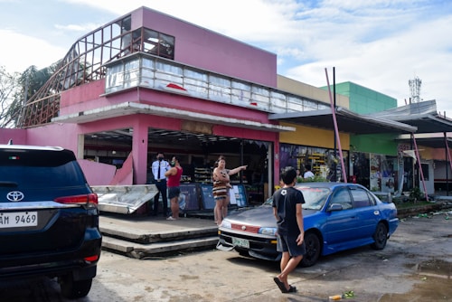 A damaged building with a pink exterior is partially destroyed, with debris scattered around. People are standing and discussing the situation, while a security guard observes. Two parked cars, one black and one blue, are visible in the foreground. The scene conveys a sense of aftermath and recovery.