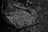 A black and white photo depicts shattered glass pieces scattered on a crumpled surface that appears to be plastic or thin fabric. The sharp fragments reflect light, creating small, bright spots against the darker background.