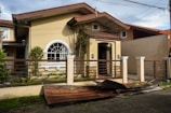 A small beige-colored house with a damaged metal roof panel in front, laying on a concrete driveway. The house features a large arched window and is surrounded by a brown fence and some greenery. The sky appears cloudy and power lines are visible above.