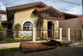 A small beige-colored house with a damaged metal roof panel in front, laying on a concrete driveway. The house features a large arched window and is surrounded by a brown fence and some greenery. The sky appears cloudy and power lines are visible above.