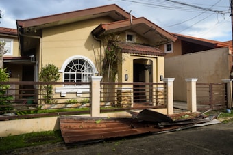 A small beige-colored house with a damaged metal roof panel in front, laying on a concrete driveway. The house features a large arched window and is surrounded by a brown fence and some greenery. The sky appears cloudy and power lines are visible above.