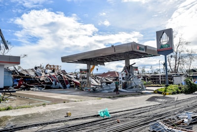 A gas station is severely damaged, with debris scattered around. The metal roof is partially collapsed, and various signs of destruction are visible. The sky is partly cloudy, and the area appears to have been affected by a natural disaster.