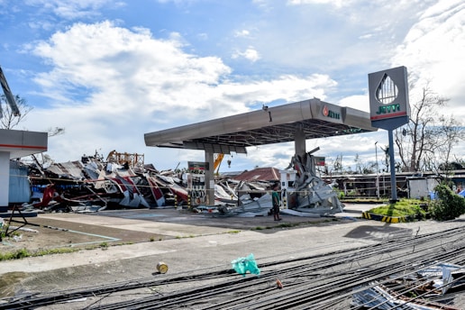 A gas station is severely damaged, with debris scattered around. The metal roof is partially collapsed, and various signs of destruction are visible. The sky is partly cloudy, and the area appears to have been affected by a natural disaster.