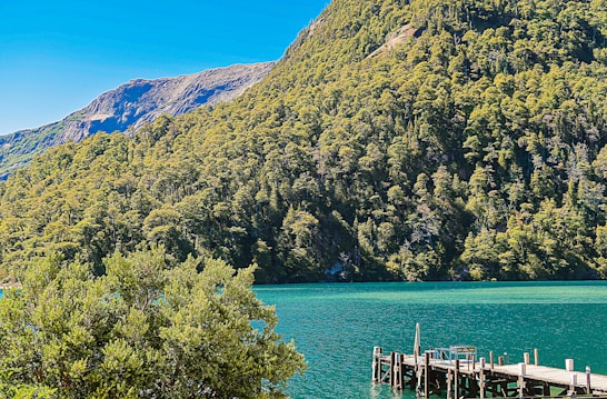 A serene landscape featuring a turquoise lake bordered by lush green forested hills under a clear blue sky. A wooden dock extends into the lake from the right side, suggesting a tranquil setting for relaxation or contemplation.