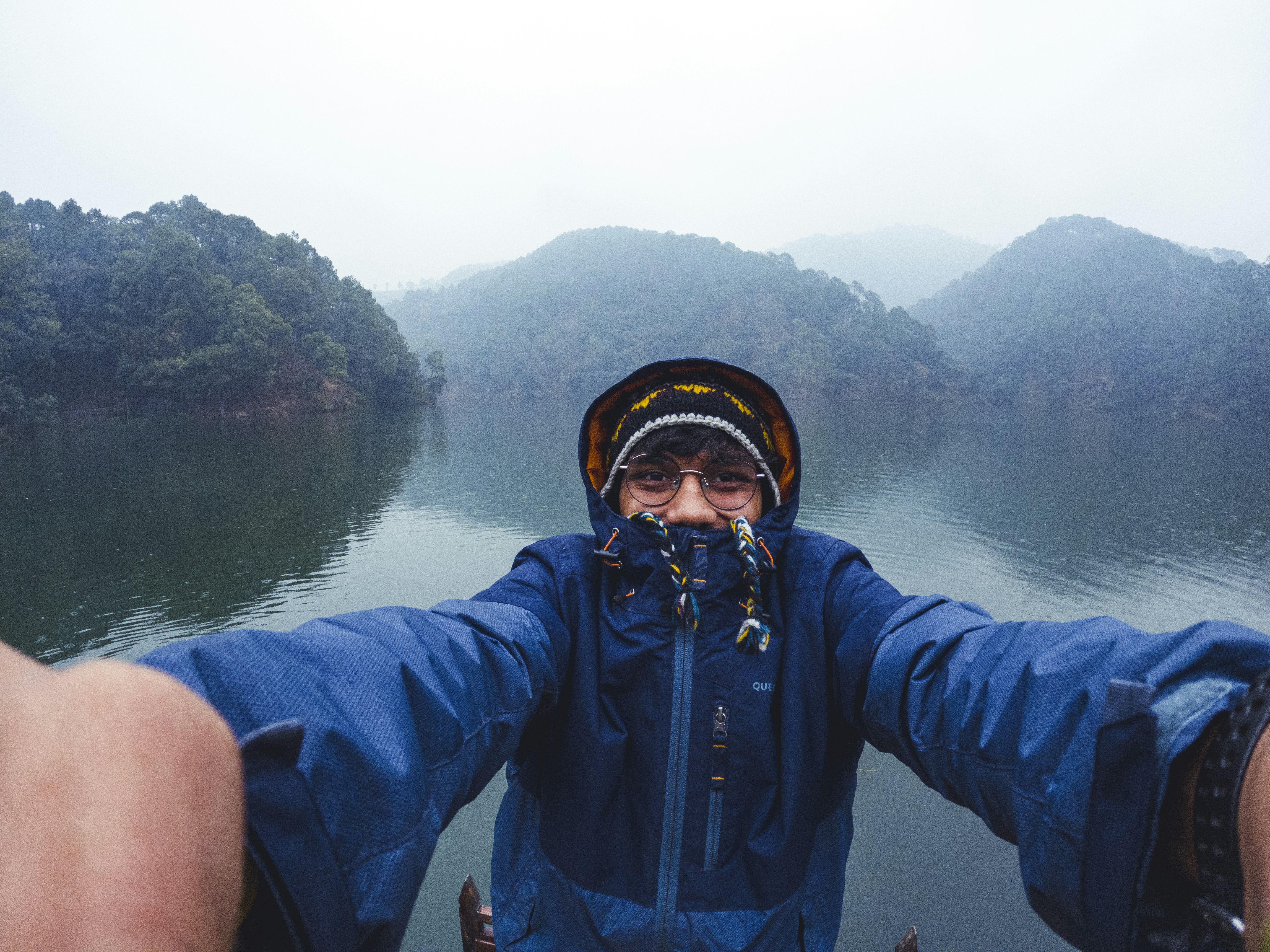 A person in a blue waterproof jacket extends arms toward the camera on a small boat, over a calm lake with forested, misty hills in the distance.