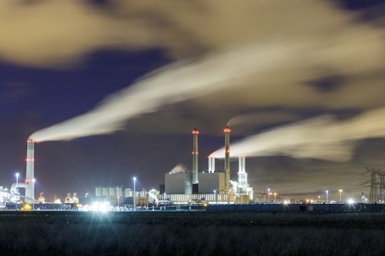 An industrial power plant is illuminated against the night sky, with smoke billowing from several tall chimneys. The facility is surrounded by lights, and clouds are visible in the background.