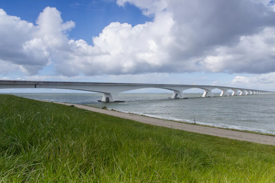 The image depicts a long, white bridge spanning across a body of water. The bridge is supported by two sturdy pillars on each side and features multiple arches that add to its architectural appeal. The perspective of the photo is from ground level, looking down the length of the bridge towards the water below. The sky above is filled with clouds, casting a soft light over the scene. On either side of the river, there are patches of grass visible on the left and right sides of the image.