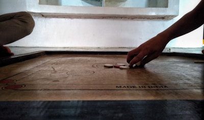 A person is playing carrom, with their hand poised to strike the striker on a wooden game board. The board is marked with a label 'MADE IN INDIA', and several carrom discs are visible. The setting appears to be indoors, with another person partially visible sitting near the board.