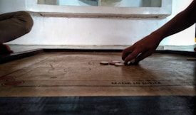 A person is playing carrom, with their hand poised to strike the striker on a wooden game board. The board is marked with a label 'MADE IN INDIA', and several carrom discs are visible. The setting appears to be indoors, with another person partially visible sitting near the board.