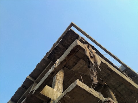 A partially constructed or dilapidated concrete structure is viewed from below against a bright blue sky. Some damaged or frayed materials hang from the concrete beams.