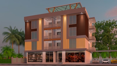 A modern four-story building with large glass windows and a rooftop terrace. The facade features warm tones, with vertical and horizontal design elements. Palm trees and other greenery surround the building, and two cars are parked on the street. The ground floor has what appears to be a commercial space with visible seating arrangements.
