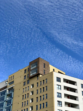 A modern residential building with beige and white tones surrounded by green spaces under a clear blue sky.