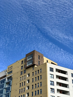 A modern multi-story residential building with a beige facade and large windows is set against a backdrop of a clear blue sky with scattered, textured clouds. The building features a mix of glass and solid materials, with balconies visible on the right side.